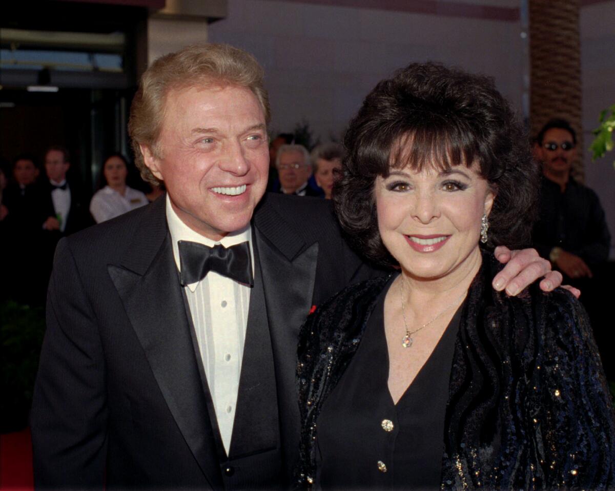 A man in a black and white tuxedo smiling to his side and standing next to a woman in a dark dress posing for photos
