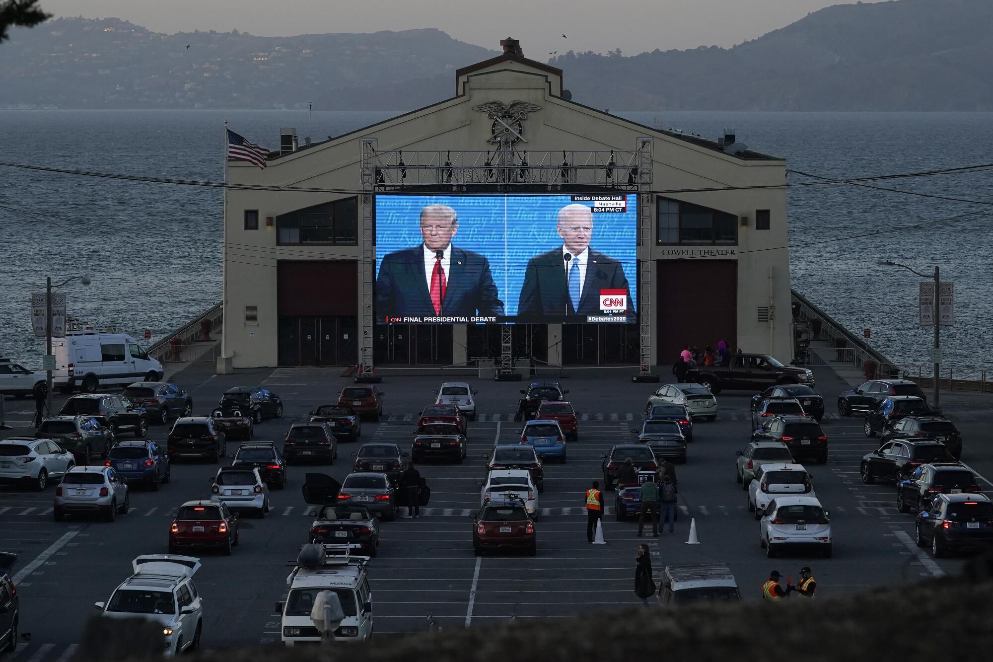 Cars parked in front of a large outdoor video screen showing the debate