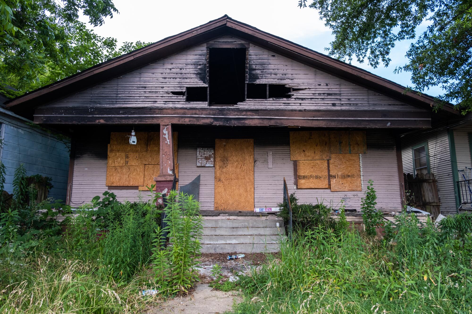 A house on an overgrown lot has a hole in the roof and boarded-up windows and door.