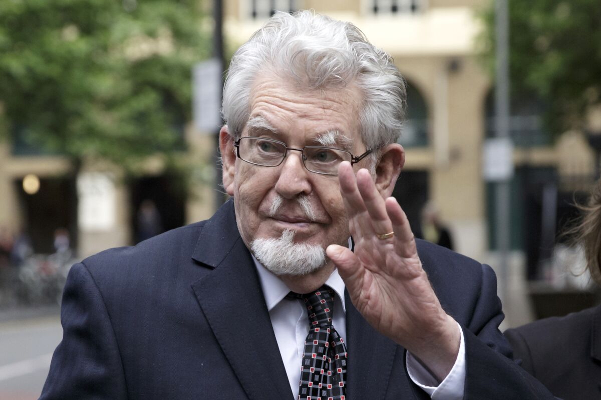 Rolf Harris waves while walking outdoors in a suit and glasses