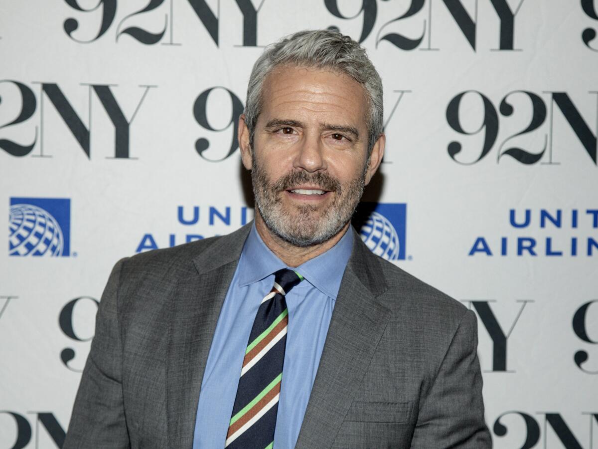 Andy Cohen poses in front of a promotional backdrop in grey suit, blue shirt and multicolor tie