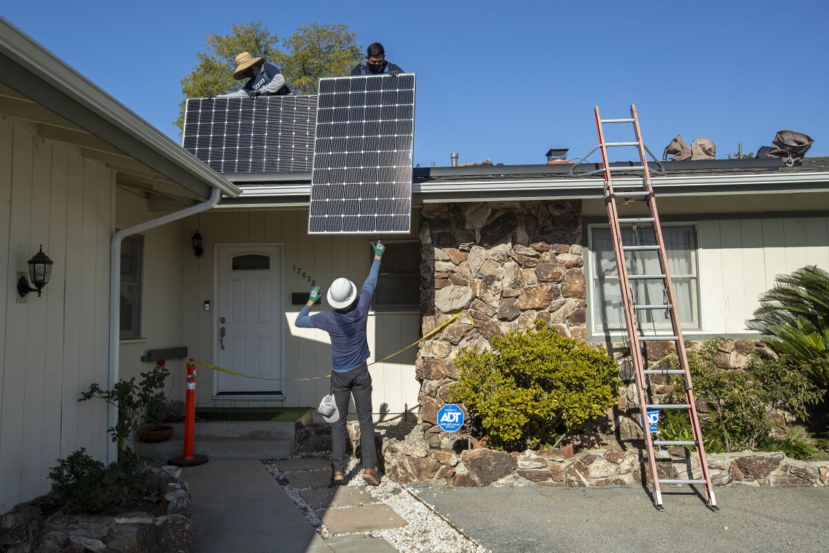 Two men on the roof of a one-story home hold solar panels; another man stands below. 