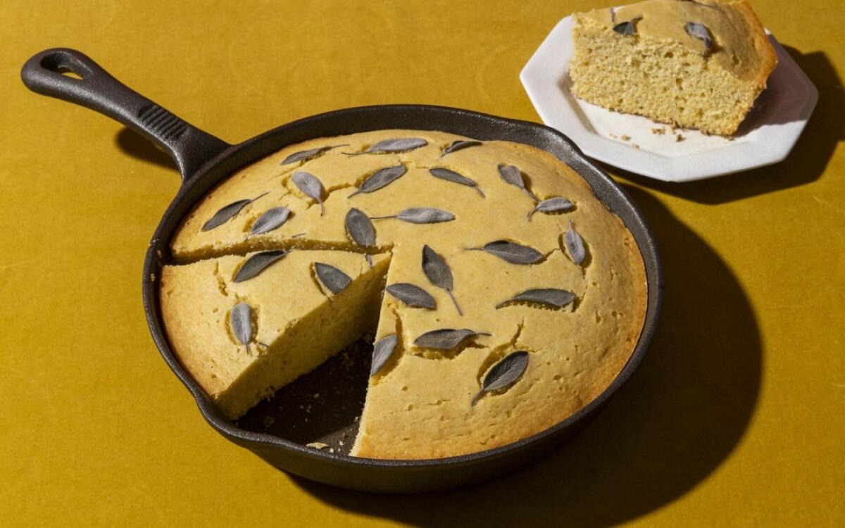 Cast-Iron Cornbread in a cast-iron skillet, garnished with sage leaves, one slice removed and sitting on a plate.