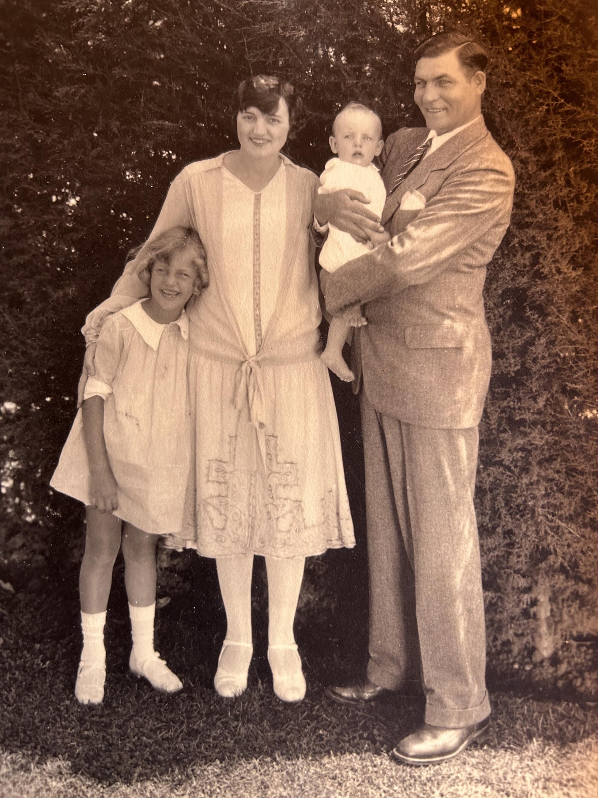 A man stands holding a baby next to a woman and a young girl, all in 1920s clothing in a vintage photo