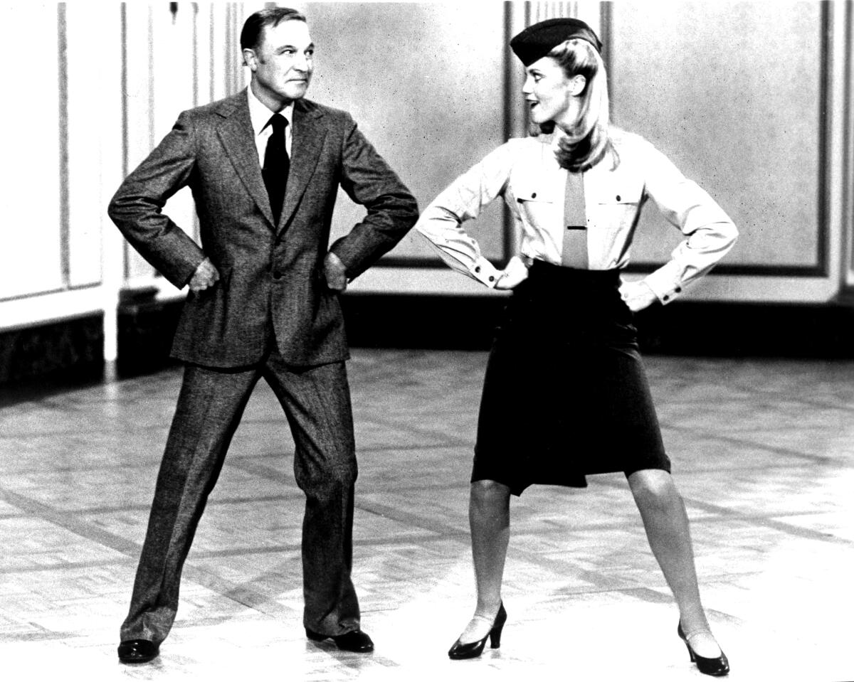 A black-and-white image of a man in a suit and a woman in a hat, jacket and skirt posing akimbo