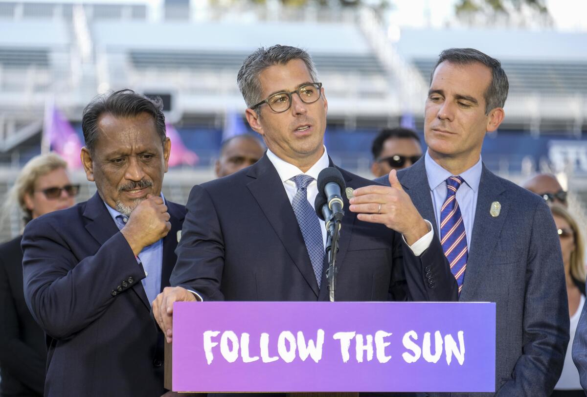 A man in a suit at a podium with a sign that says "Follow the Sun," flanked by two other men in suits