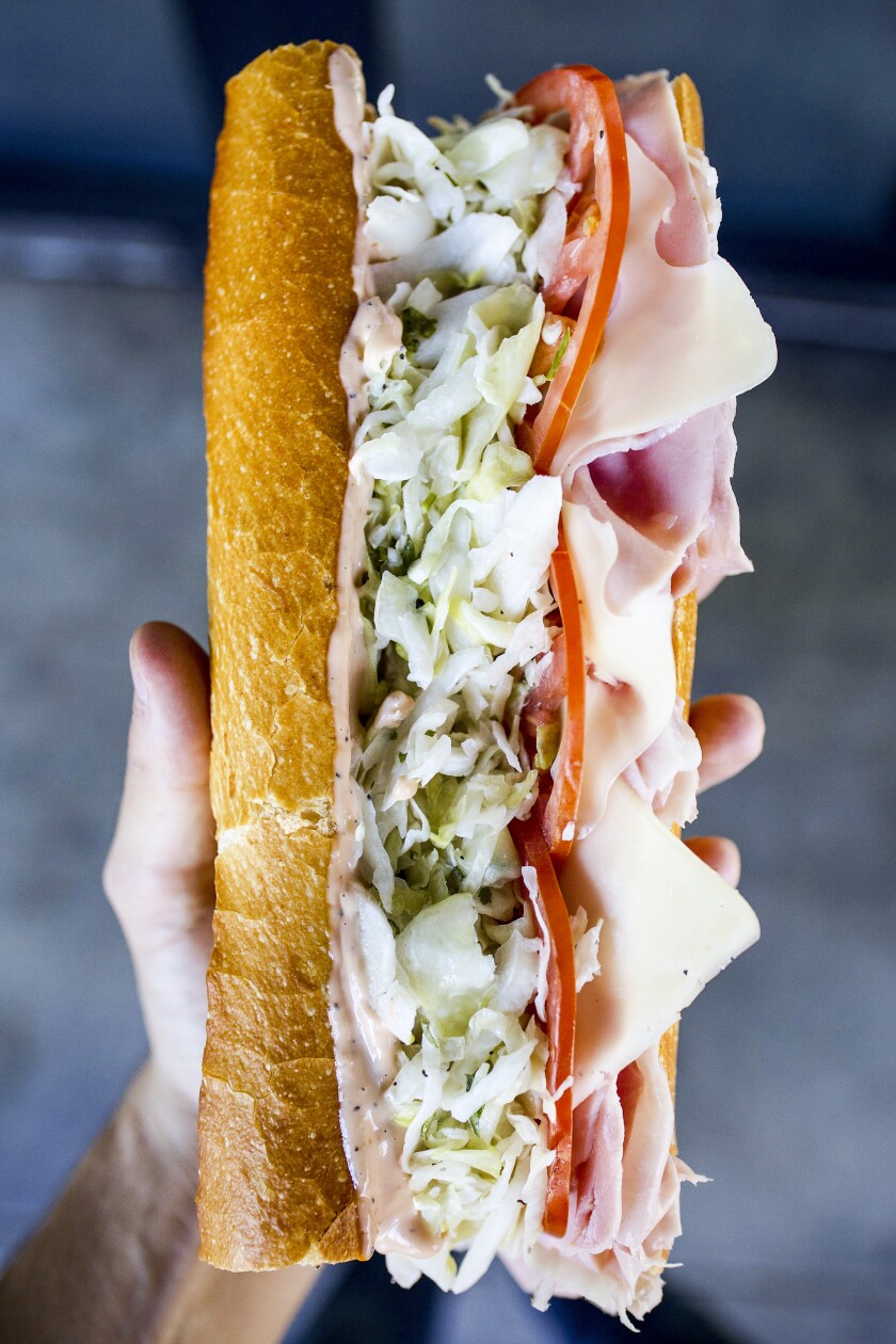 Ein Foto einer Hand, die ein großes Schinken-Käse-Sandwich auf Baguette hält, der Heavenly Ham N Swiss.