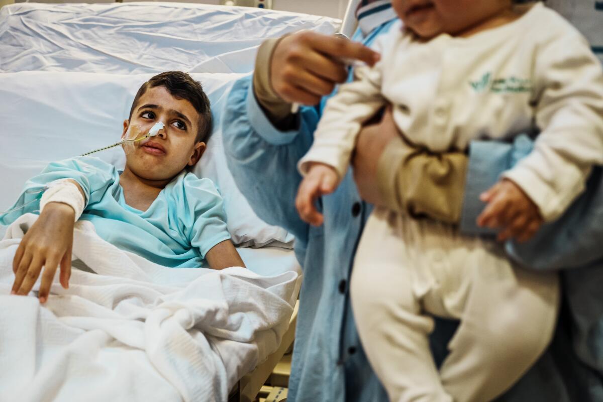 一名 10 岁的巴勒斯坦男孩躺在医院的病床上，前景中他的母亲抱着一个婴儿。 