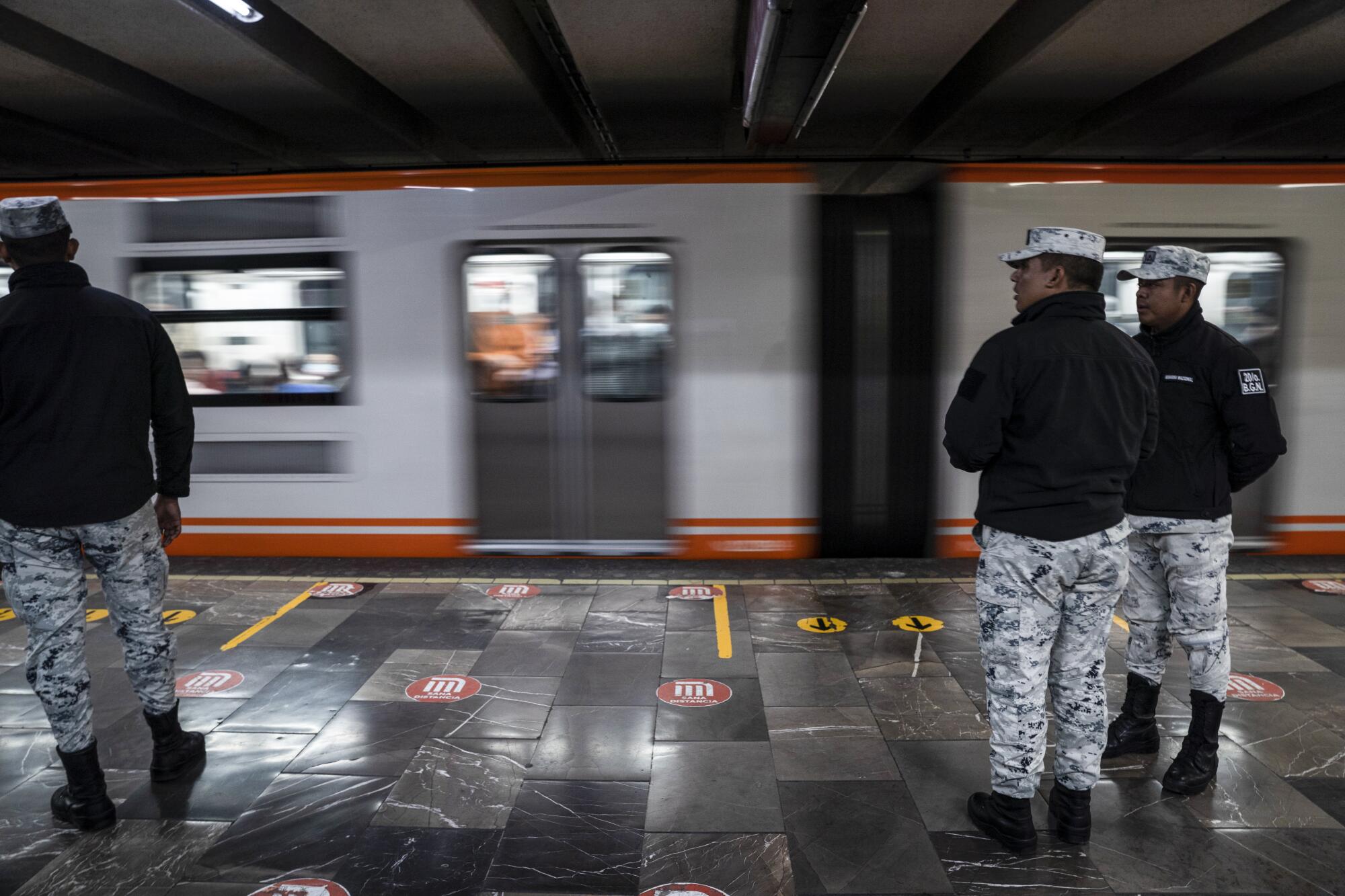 Three people in identical outfits stand on a platform while a subway train speeds by.