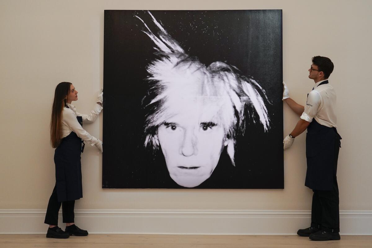 A woman and a man stand on either side of a large self-portrait by Andy Warhol with his face against a black background.
