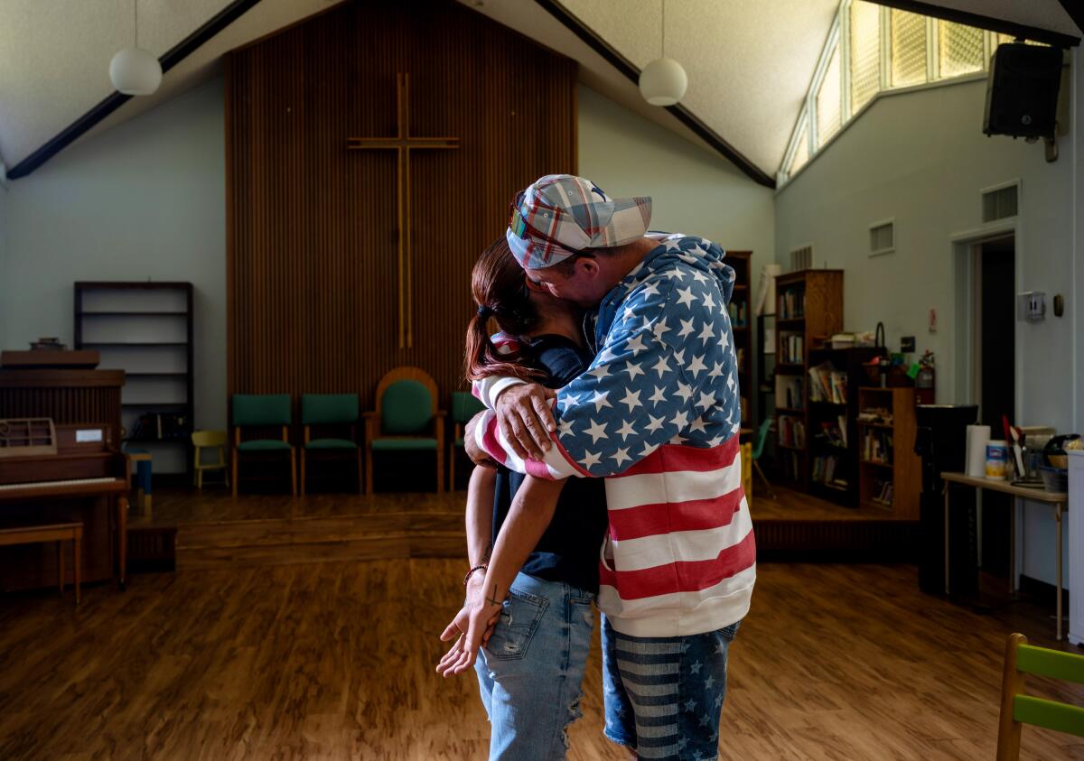 A man an woman embrace in a church. The man wears a stars-and-stripes shirt.
