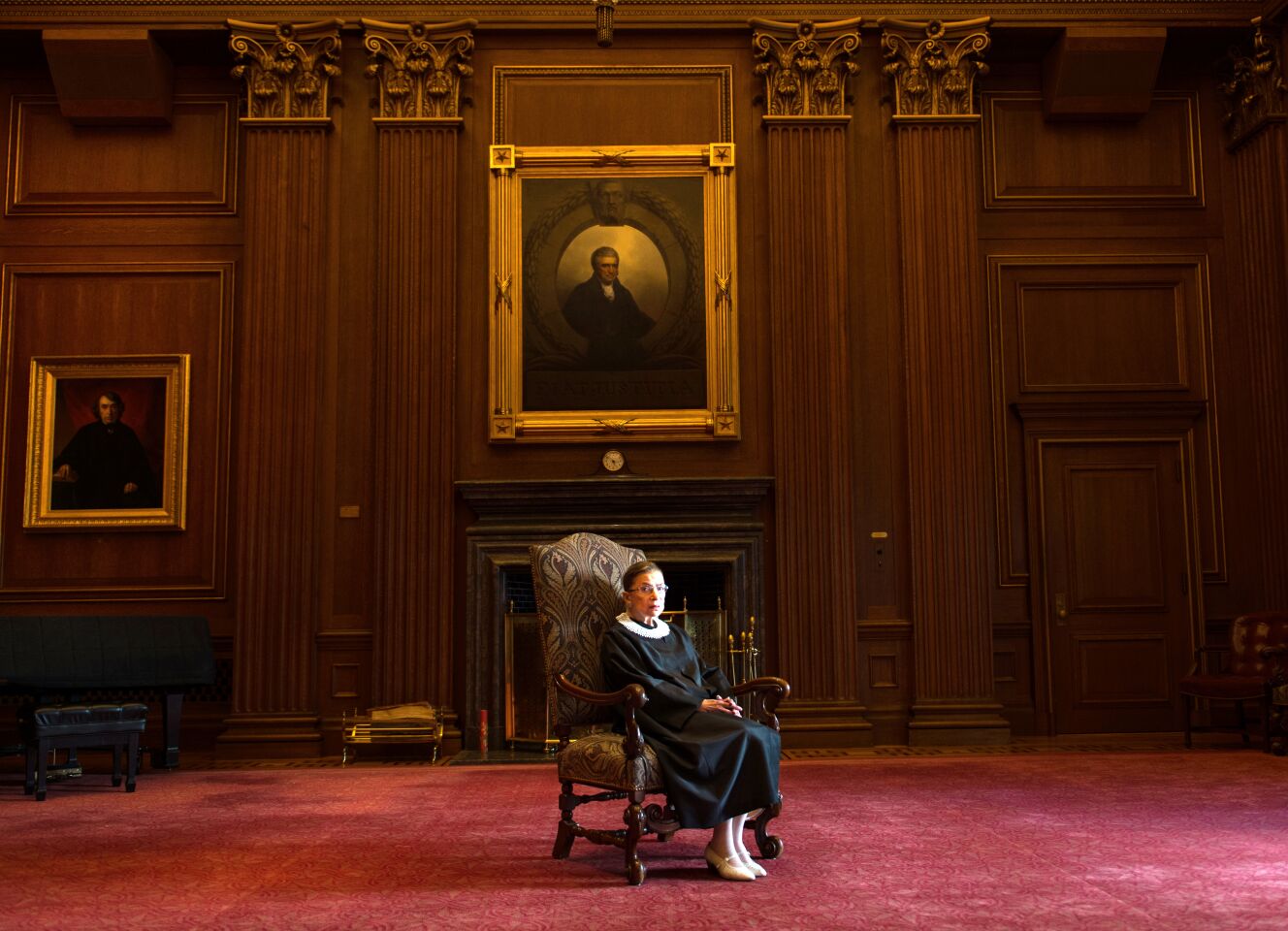 Ruth Bader Ginsburg sits in a large chair in a wood-paneled, red-carpeted room at the Supreme Court