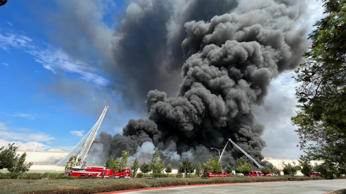 A plume of black smoke rises from a warehouse building with fire trucks outside