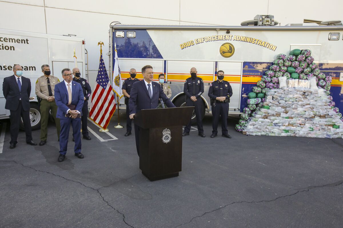 A man speaks at a lectern in front of law enforcement vehicles and officers and a large stack of plastic-wrapped packages