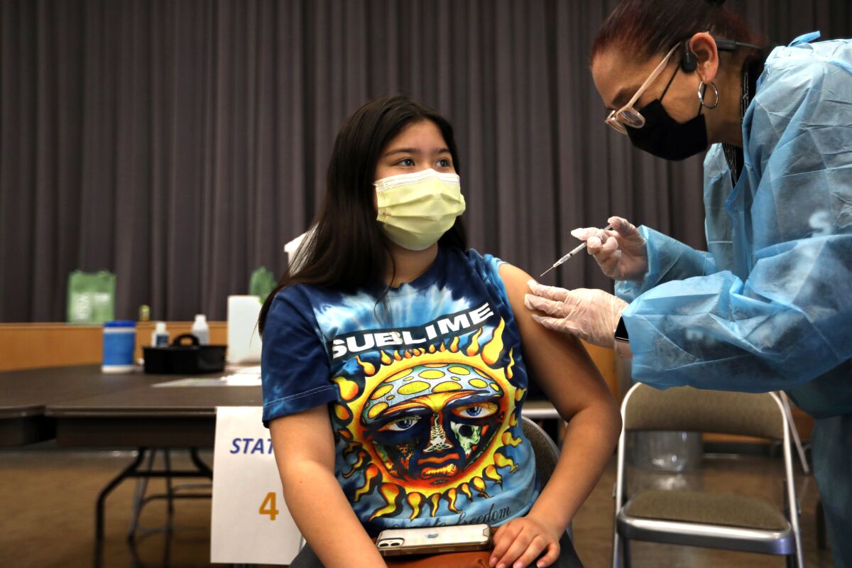 A young woman in a mask sits while receiving a shot from a health worker in protective gear.
