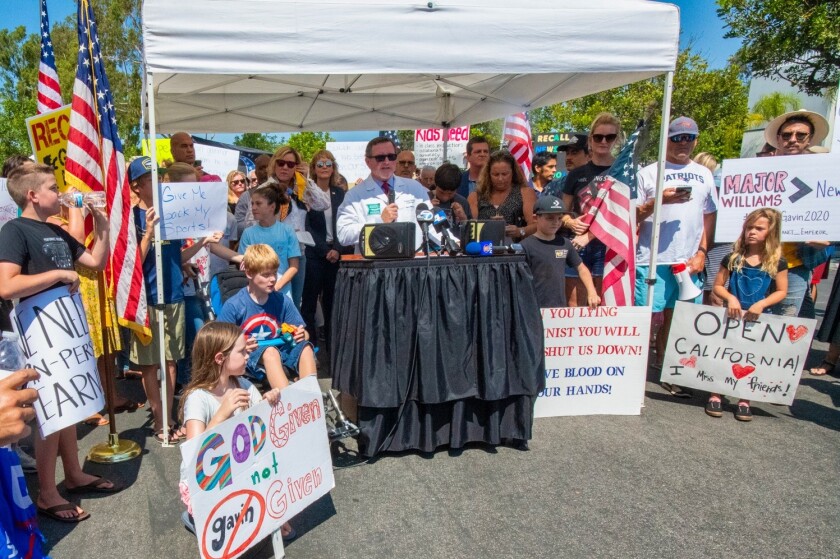 A man stands at a lectern beneath a shade canopy surrounded by adults and children holding signs calling for schools to open