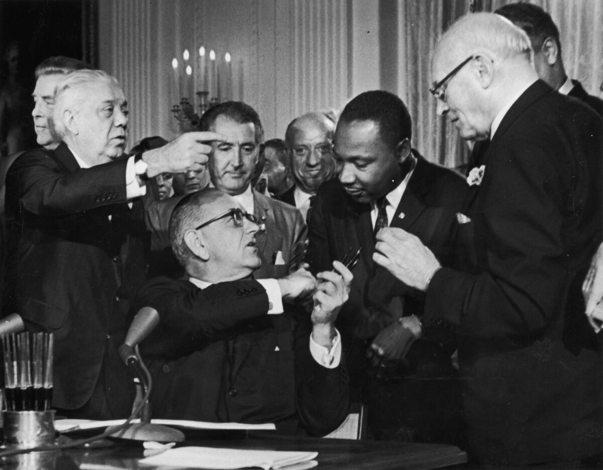 President Lyndon B. Johnson shakes the hand of Dr. Martin Luther King Jr. at the signing of the Civil Rights Act while officials look on.