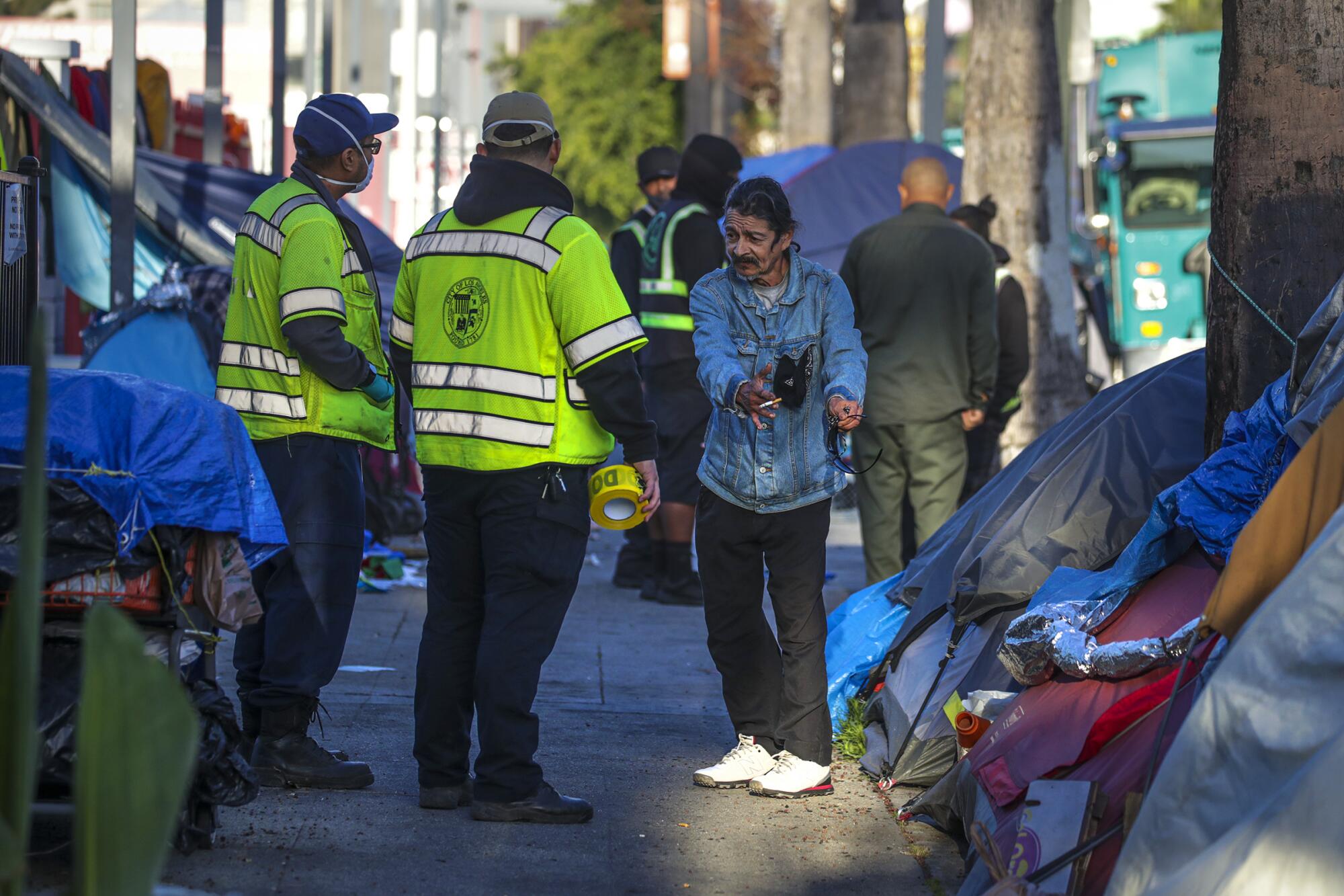 A man in a denim jacket points to his tent as he talks with city workers in yellow vests.