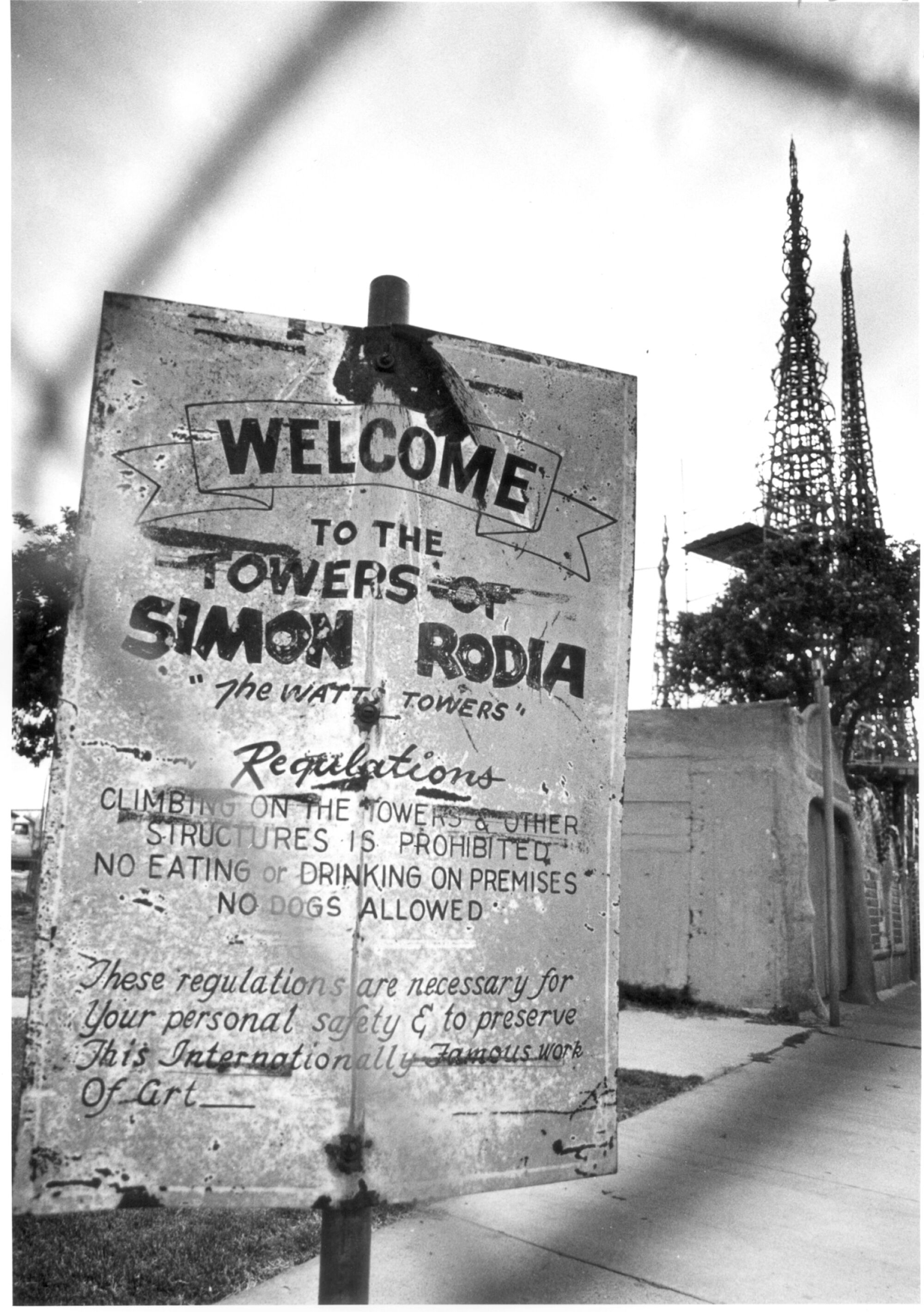 A rusted sign reading "Welcome to the Towers of Simon Rodia" with metal towers in the background