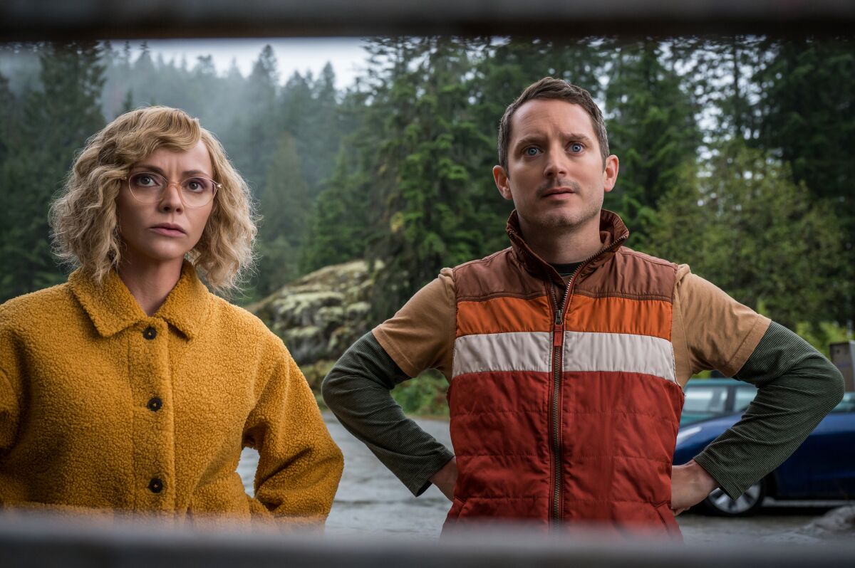 Actors Christina Ricci and Elijah Wood with their hands on their hips, with pine trees in the background