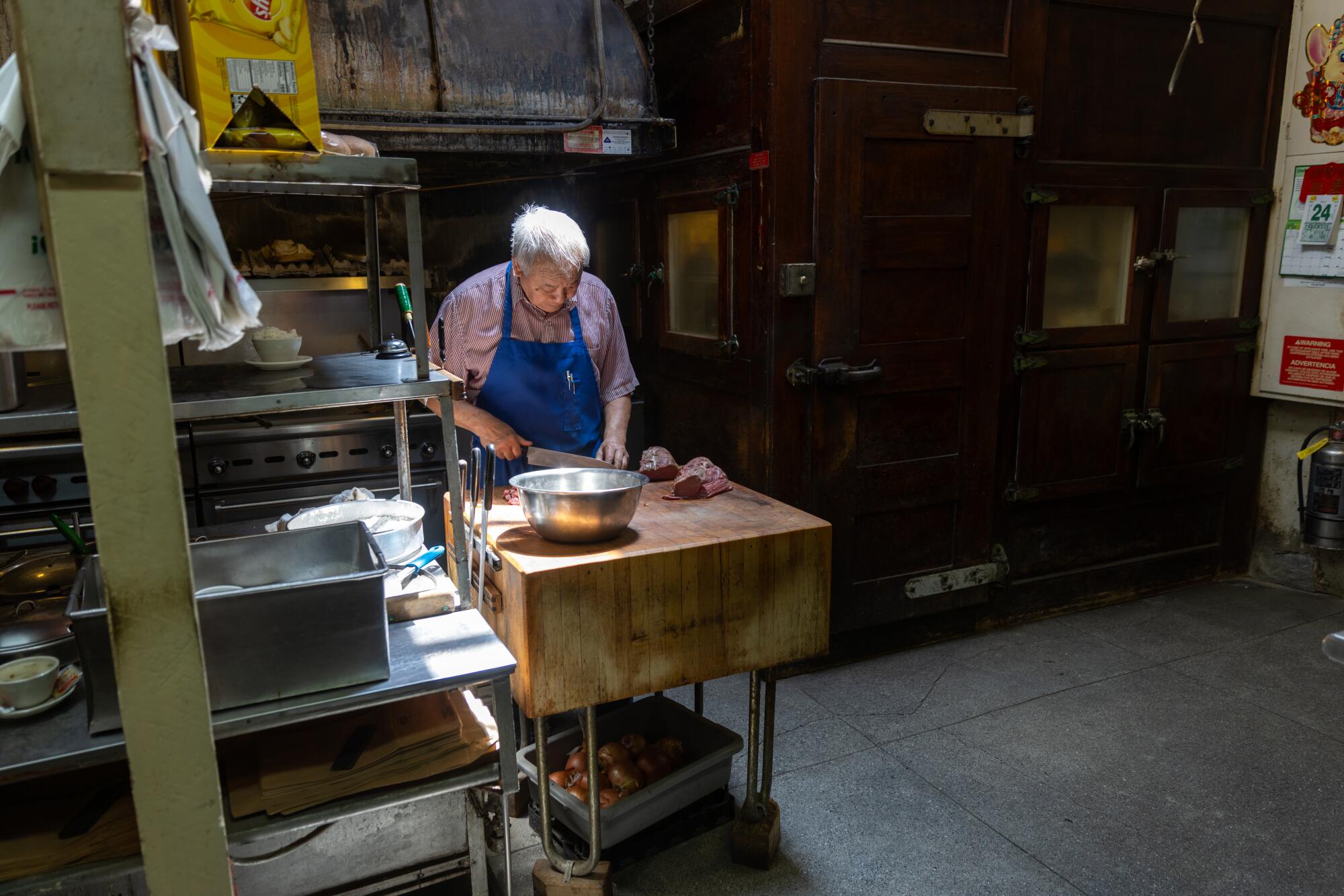 A gray-haired man in blue apron cuts meat on a butcher's block. 