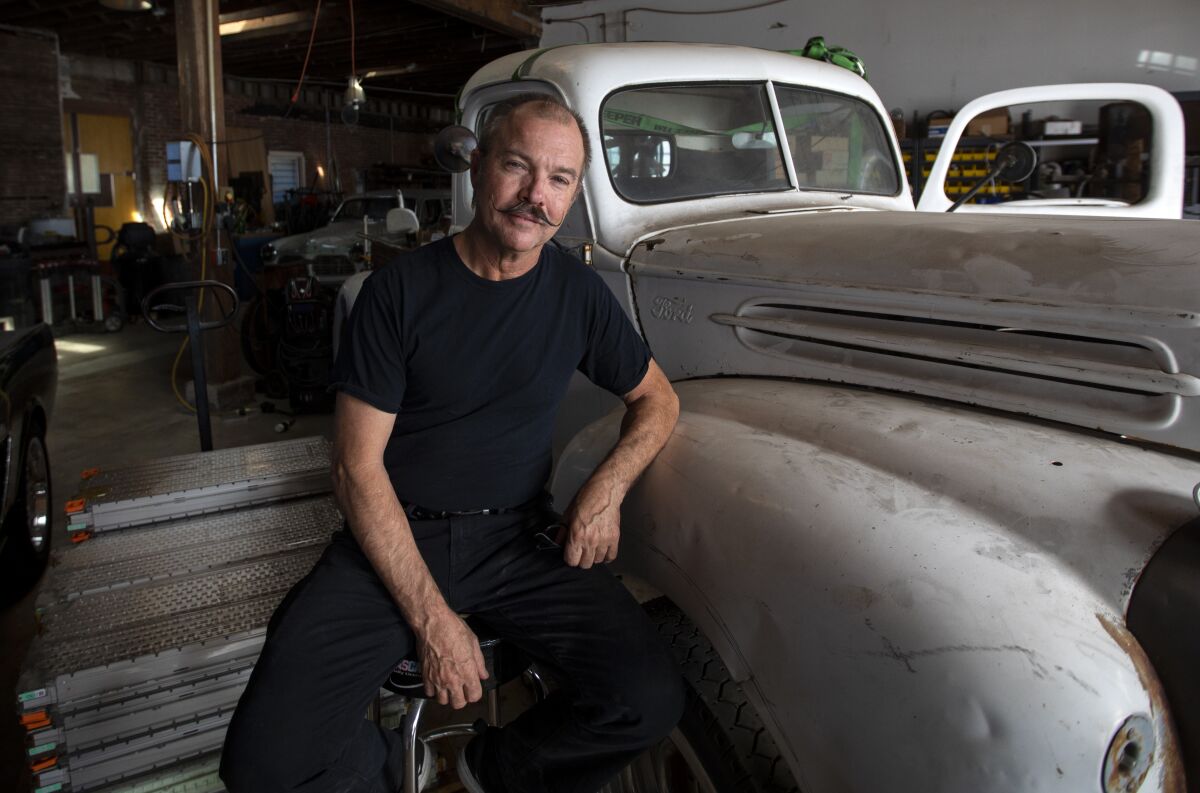 Greg "Reverend Gadget" Abbott, car inventor and EV whiz, in his shop in Los Angeles. He converts retro sports cars to electric power. He is sitting next to a 1947 Ford truck and Tesla car batteries.