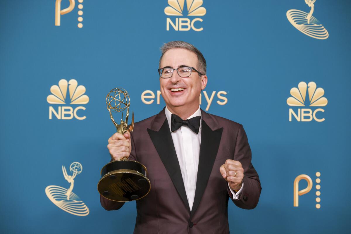 A man in a black suit, bowtie and glasses smiling and holding an Emmys trophy