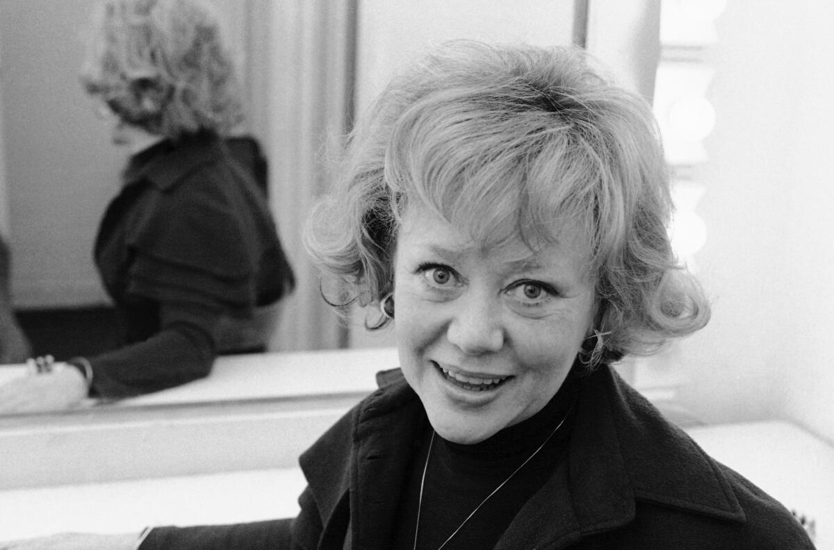 Glynis Johns wears a black coat and blouse as she poses for pictures while seated in front of a mirror