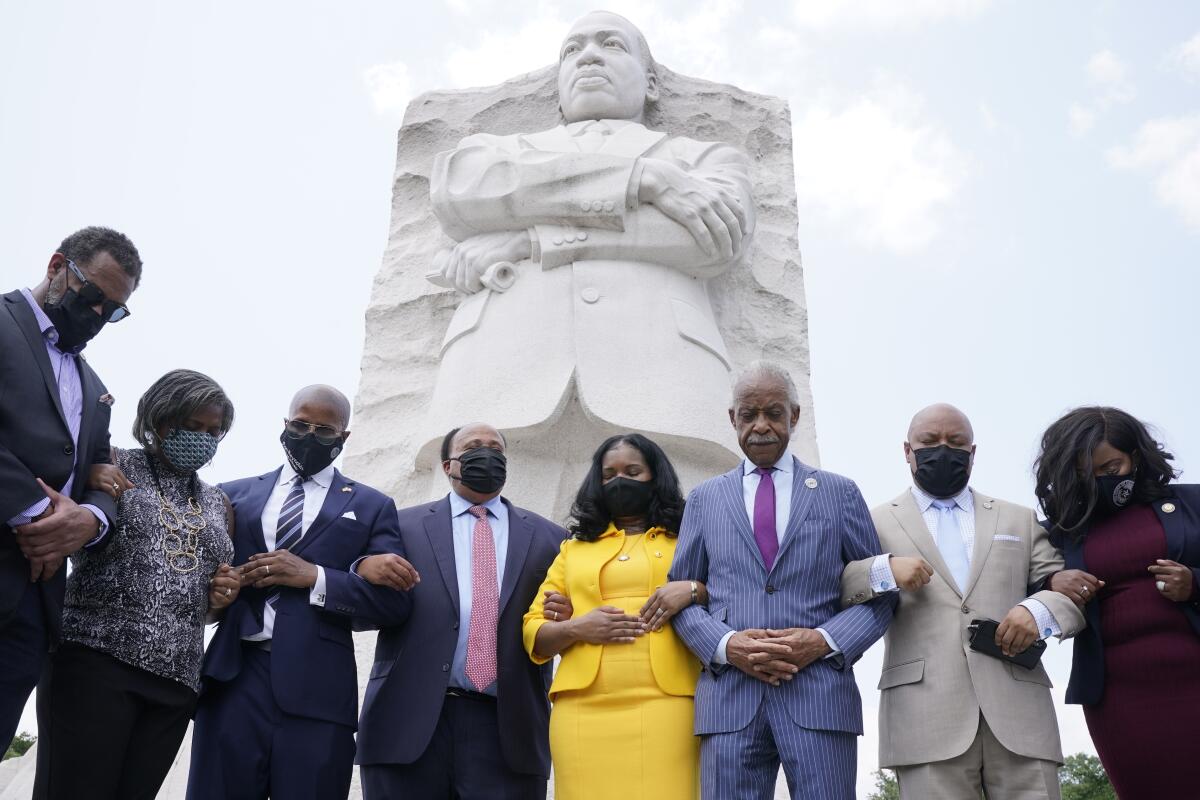 Eight people standing arm in arm with heads bowed, a large sculpture of Martin Luther King Jr. behind them.