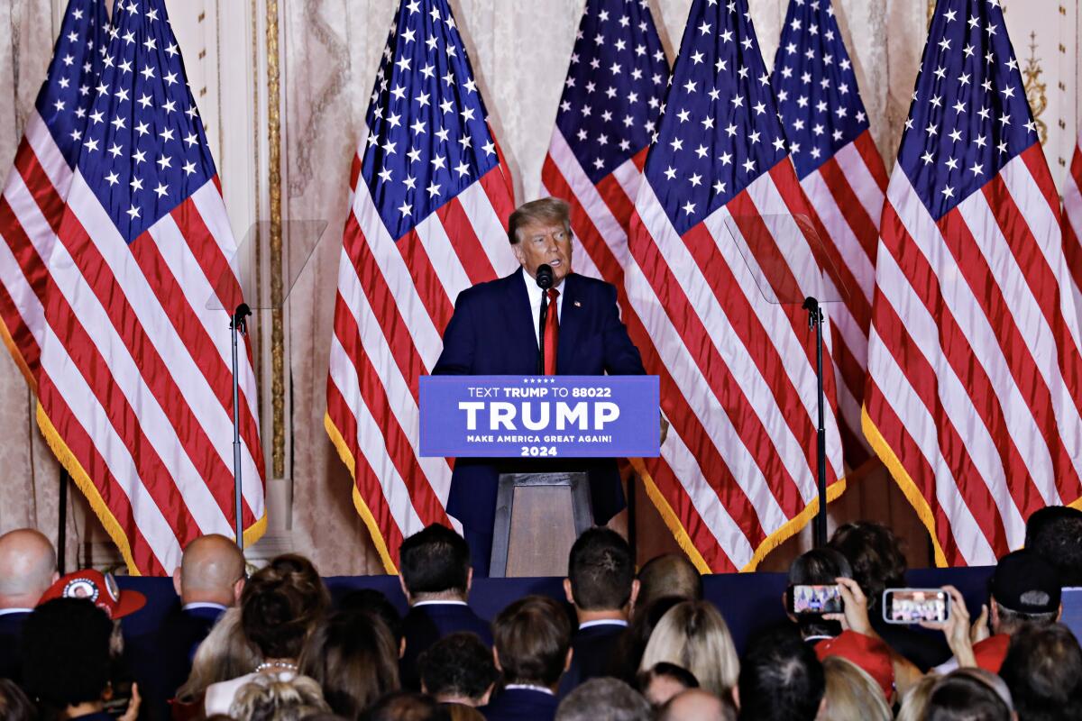 Former President Trump at a lectern bearing his name, with American flags behind him and supporters' heads in the foreground