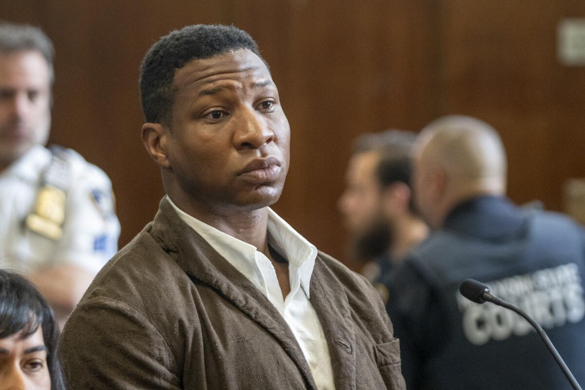 Jonathan Majors in a courtroom with a serious face while wearing a brown coat and white shirt.