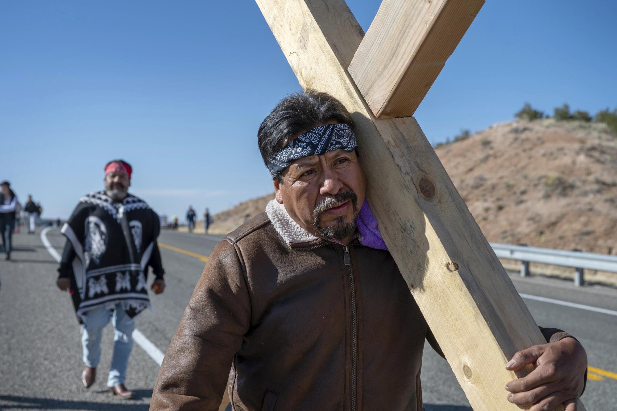 Two men walk along a highway, one of them caring a large wooden cross on his shoulder