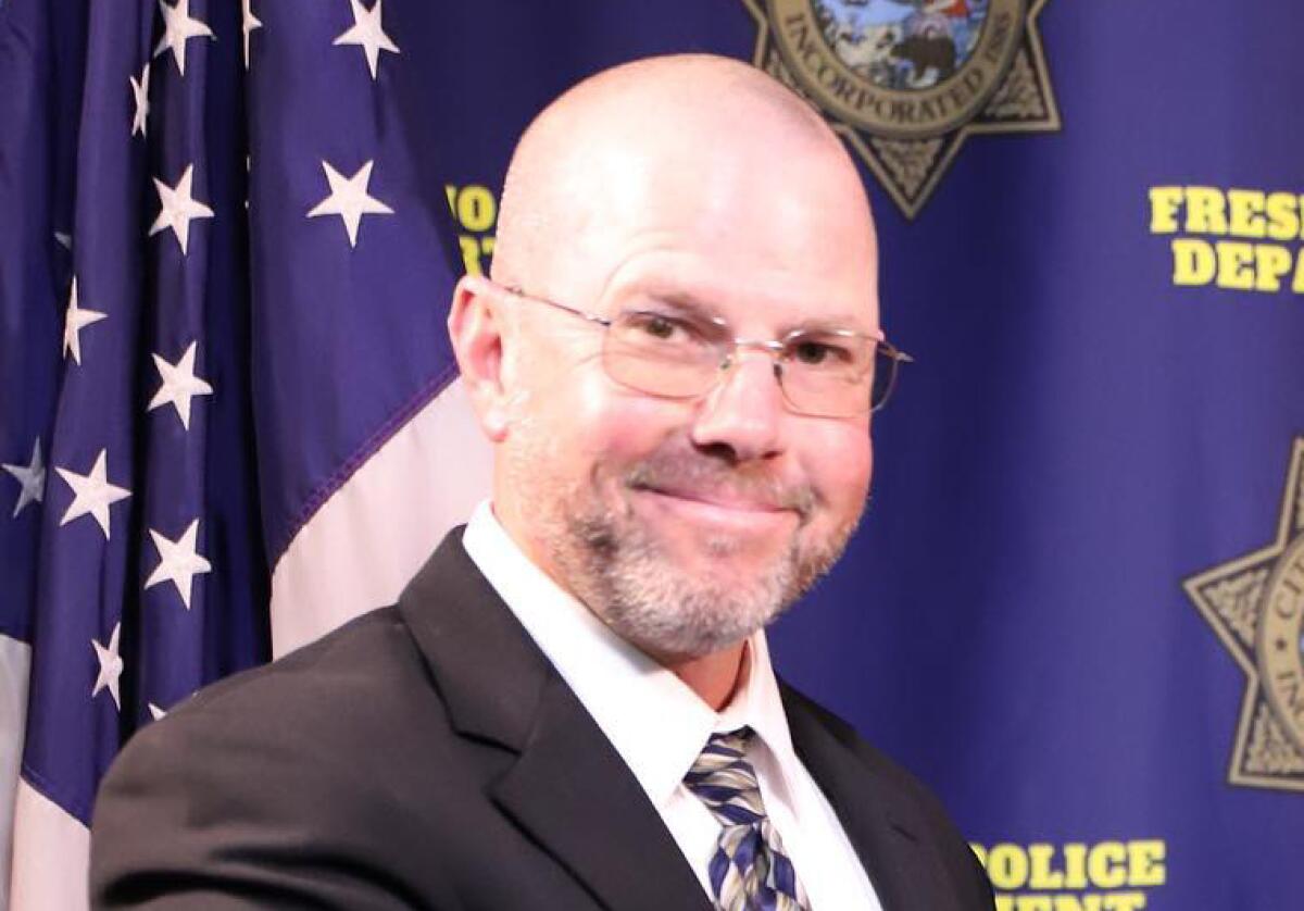 A man in glasses stands in front of a flag and a blue background.