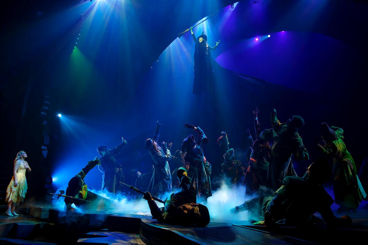 A witch rises over a stage of dancers.