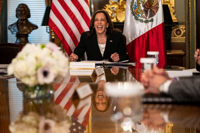 Vice President Kamala Harris laughs from the head of a conference table, with a backdrop of American and Mexican flags.