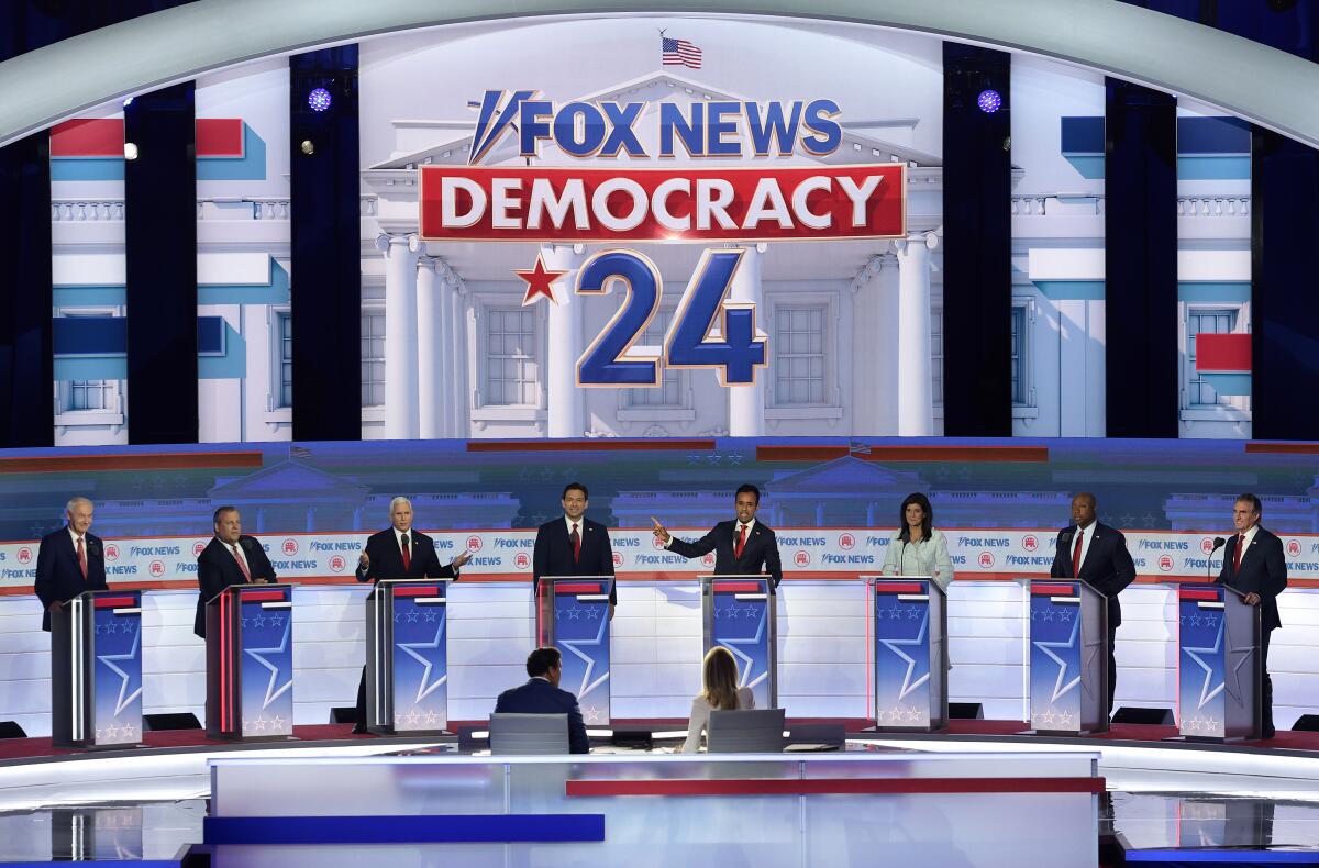 Seven men in black suits and red ties and one woman stand at podiums on stage during a presidential debate. 