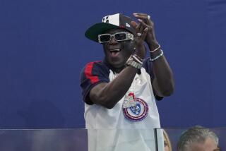 Flavor Flav smiling and clapping wearing a t-shirt and 'Team USA' hat. 