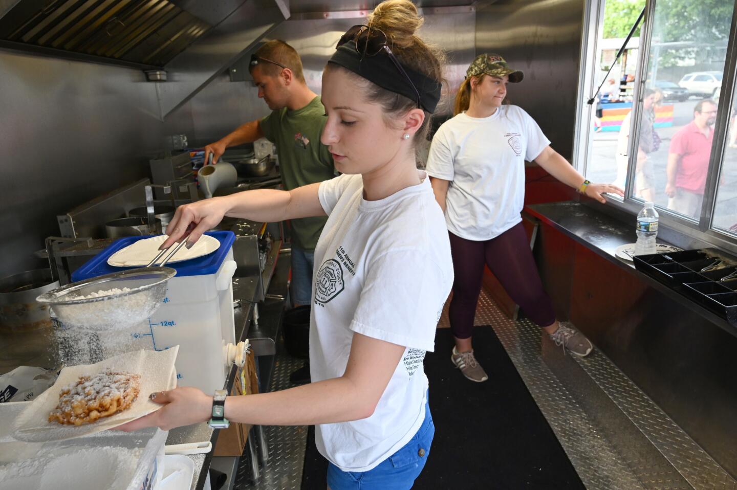 Kayla Wagerman, center, powders a funnel cake as she joined by fellow volunteers Kendall Bowers, left, and Berna Blocher, serving food from a concessions truck during the carnival at the Harney Volunteer Fire Company on Tuesday, June 25.