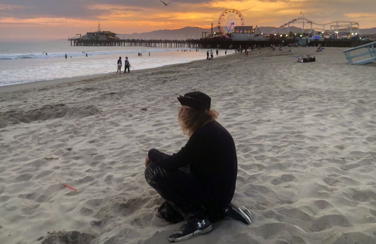 A man sits in the sand with the pier and beach in the background at sunset
