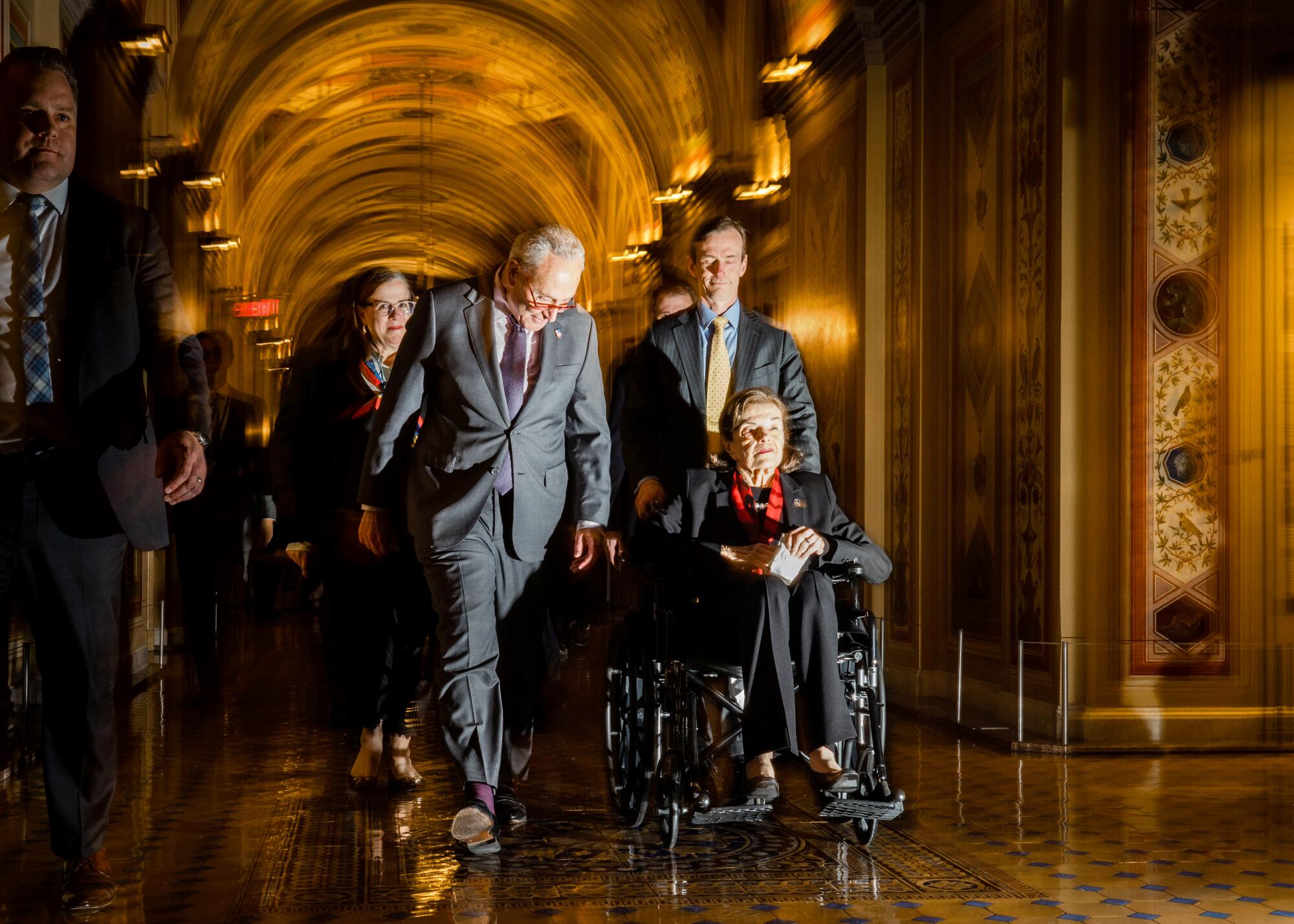 A person pushes another person in a wheelchair in an ornate hallway as others walk nearby.