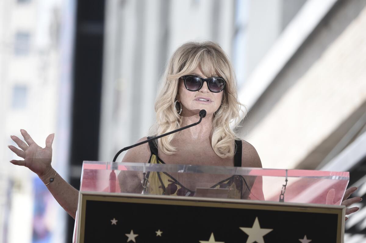 Goldie Hawn has curled blond hair and black shades on as she stands at a podium speaking. 