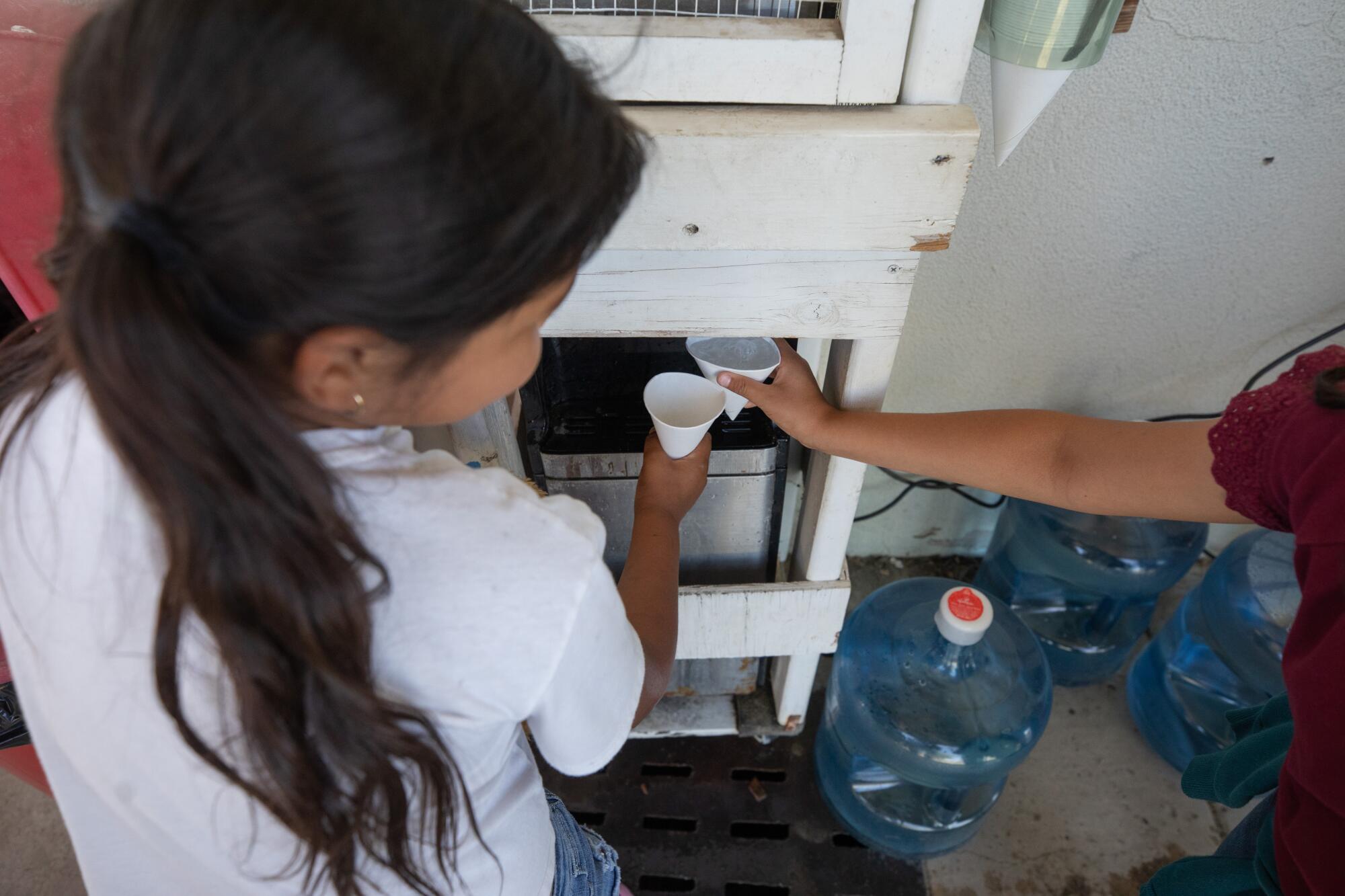 Two children fill paper cups from a water dispenser.