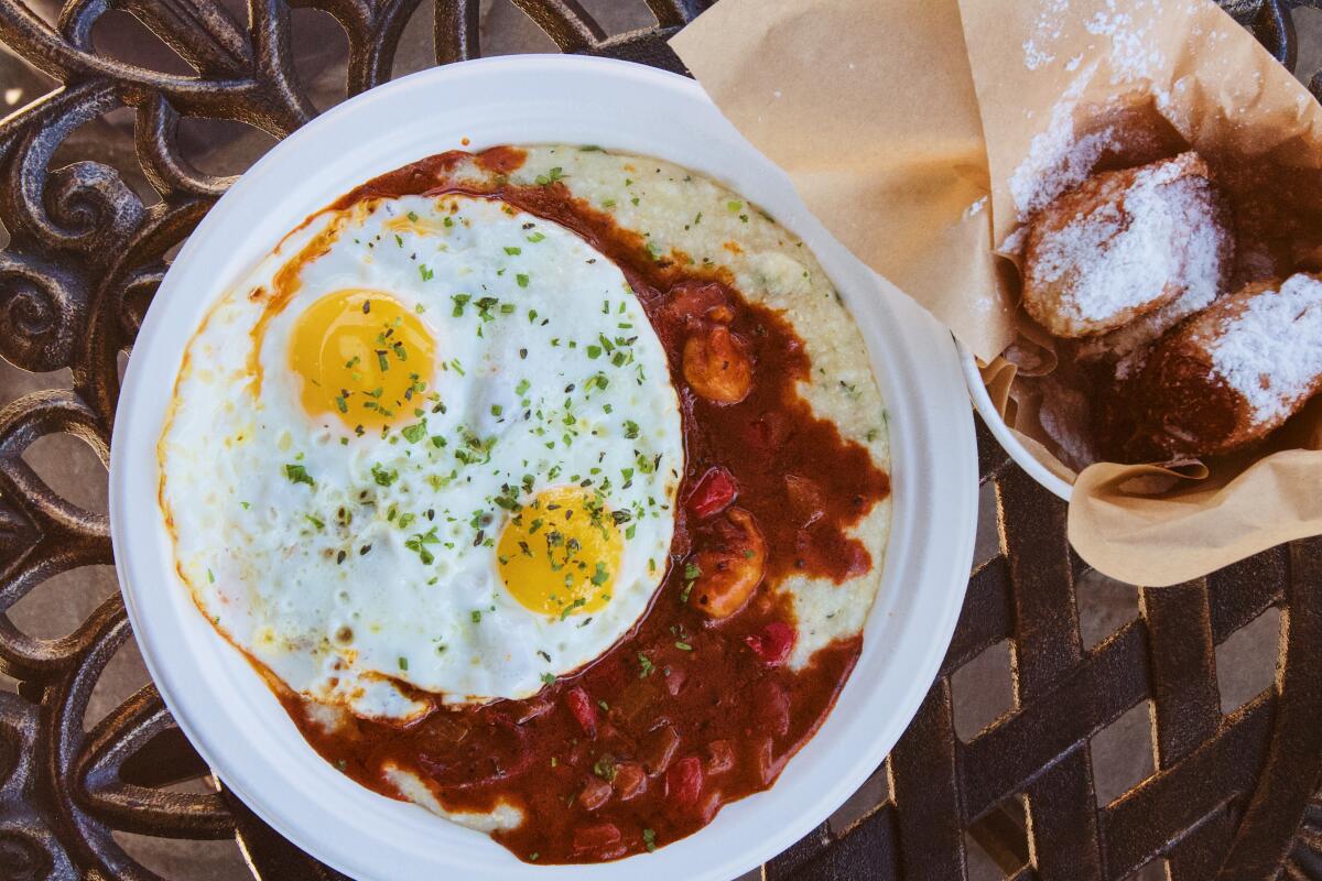 An overhead photo of a plate of shrimp and grits topped by two sunny-side-up eggs, alongside a cup of beignets.