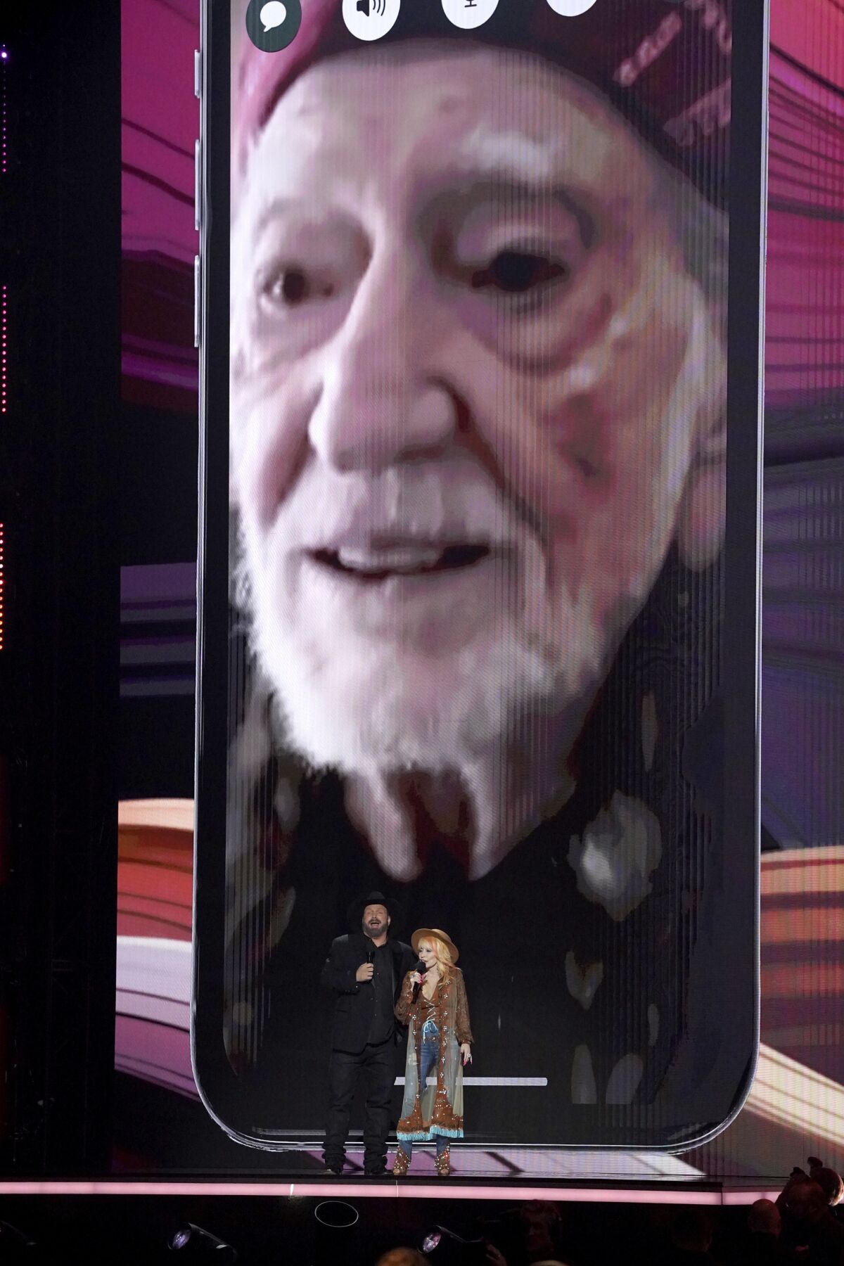 Garth Brooks and Dolly Parton stand in front of Willie Nelson on a projected screen behind them
