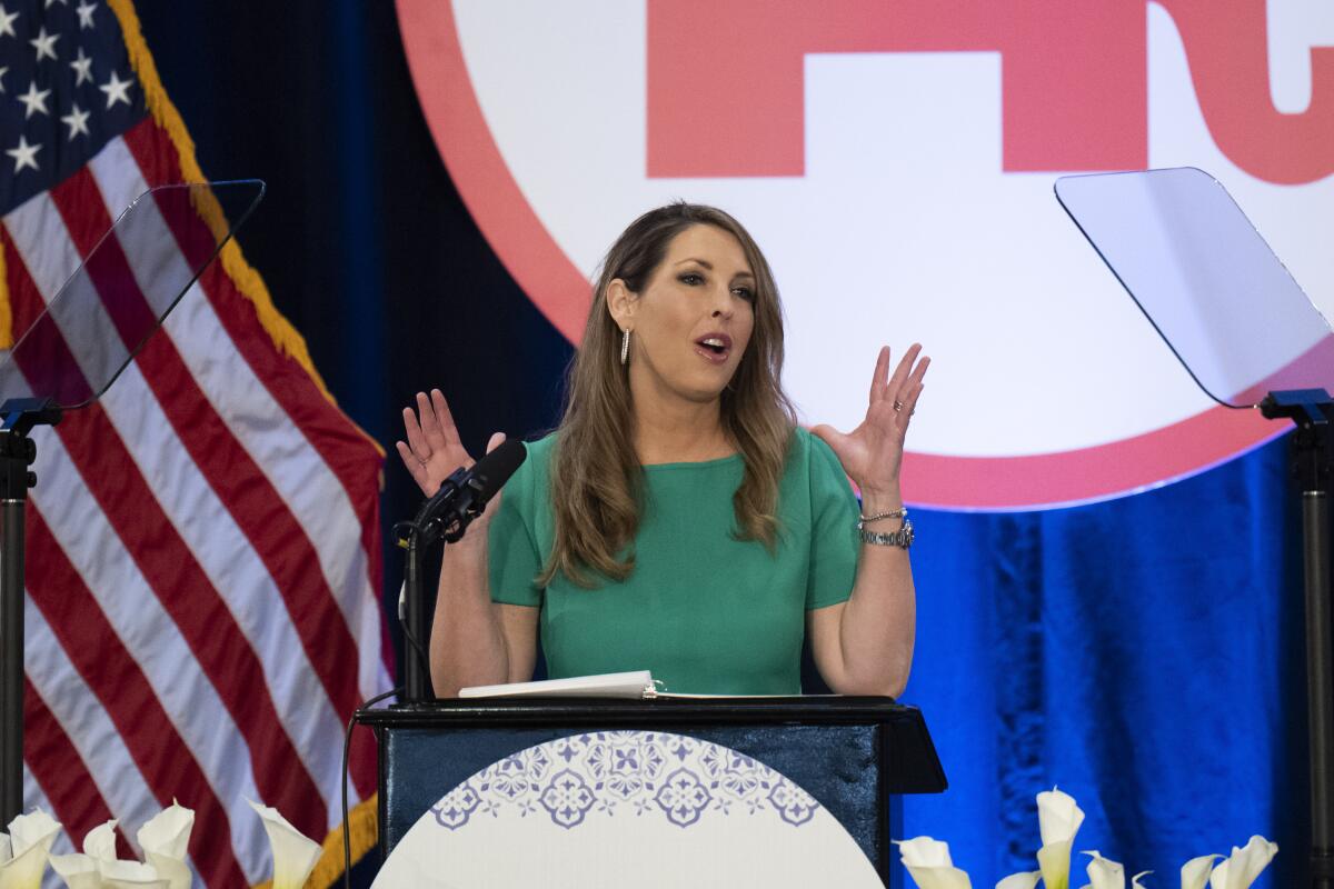Ronna McDaniel raises her hands while speaking from a lectern in front of an American flag and red, white and blue decor