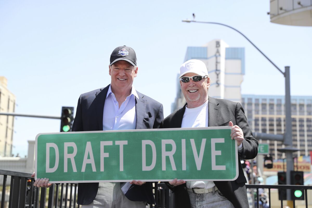 Two men sit side by side holding a street sign reading "Draft Drive"