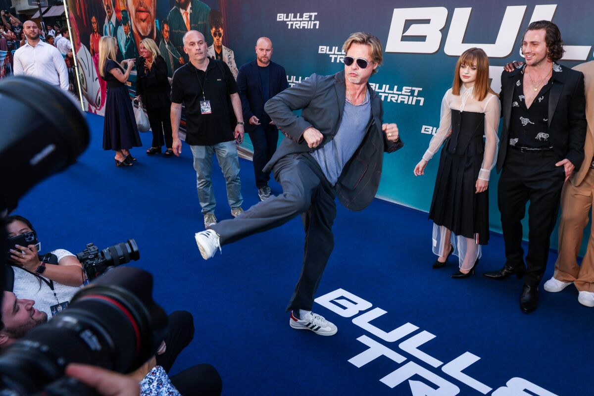 A man at a premiere in a casual blue suit and tennies karate-kicks toward photographers