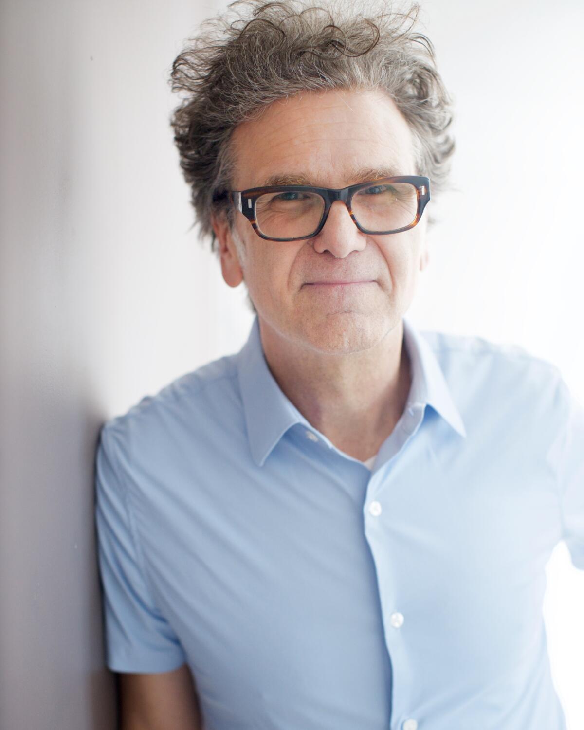 A man with wild hair wears dark-rimmed glasses and a blue shirt and leans against a wall.