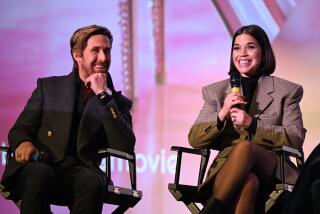 Ryan Gosling and America Ferrera sit in directors chairs onstage and smile while Ferrera speaks into a microphone