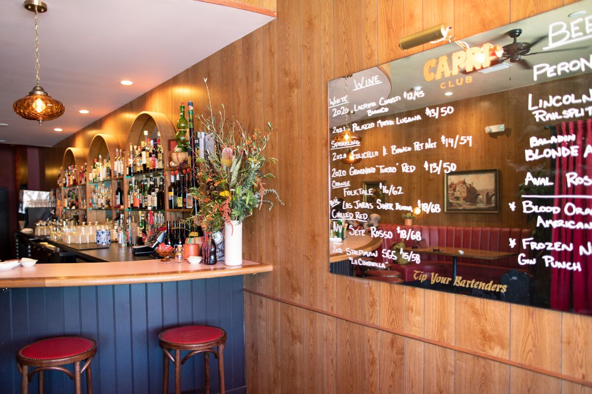 A mirror with a drinks menu written on it hangs on a wood-paneled wall in a bar.
